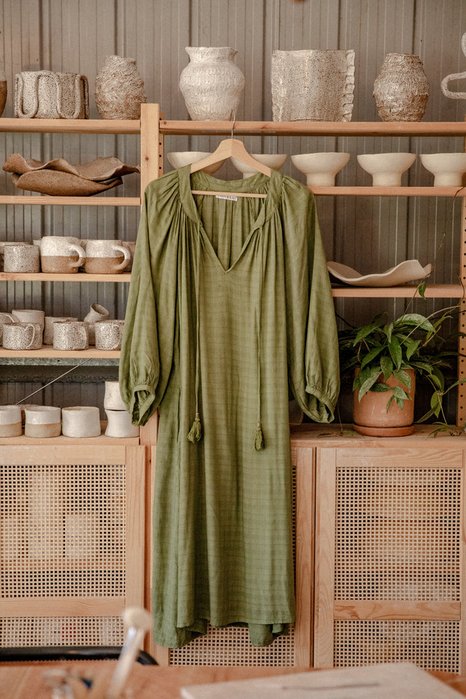 
                  
                    flores organic sugarcane dress in forest green hanging in pottery studio with ceramics in background
                  
                