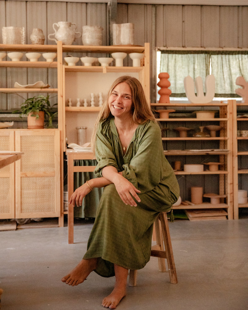 
                  
                    woman dressed in flores organic sugarcane dress in forest green sitting in pottery studio with ceramics in background
                  
                