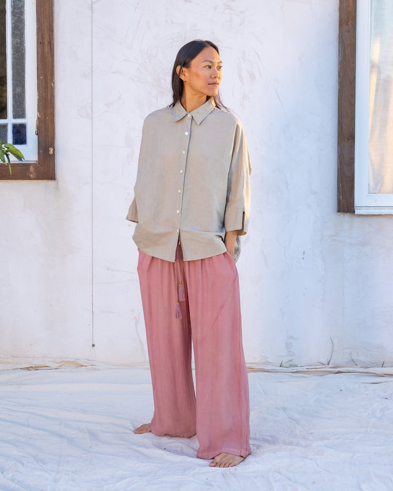 
                  
                    woman dressed in miguel straight legged pants in orange peel fibre in dirty rose pink and bouviet oversized shirt in natural linen standing in front of a white wall
                  
                