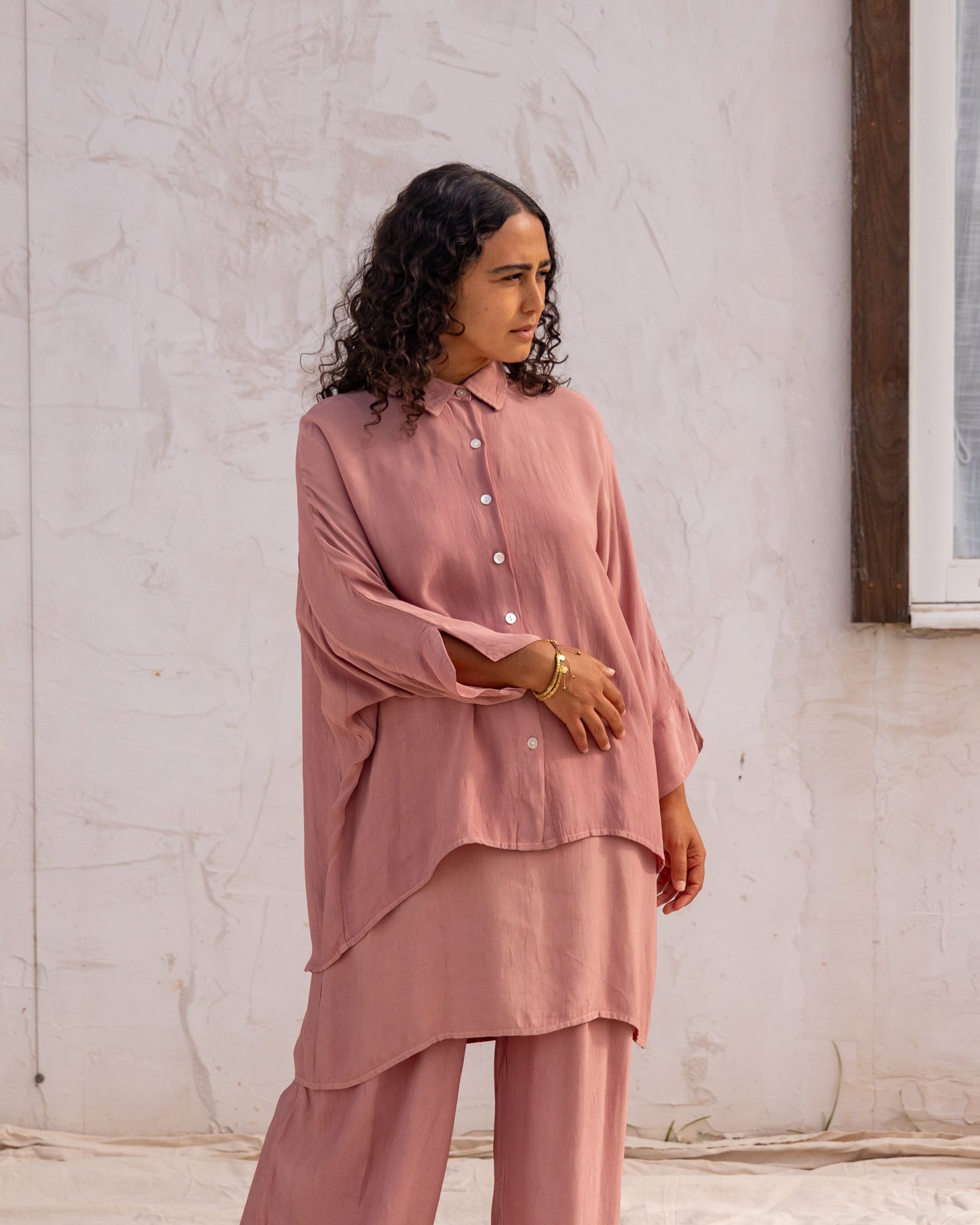 
                  
                    woman dressed in bouviet oversized shirt and miguel straight legged pants in orange peel fibre in dirty rose pink standing in front of a white wall
                  
                