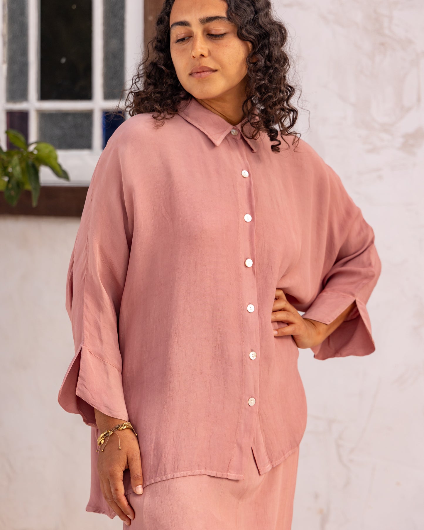 
                  
                    woman dressed in bouviet oversized shirt and miguel straight legged pants in orange peel fibre in dirty rose pink standing in front of a white wall
                  
                