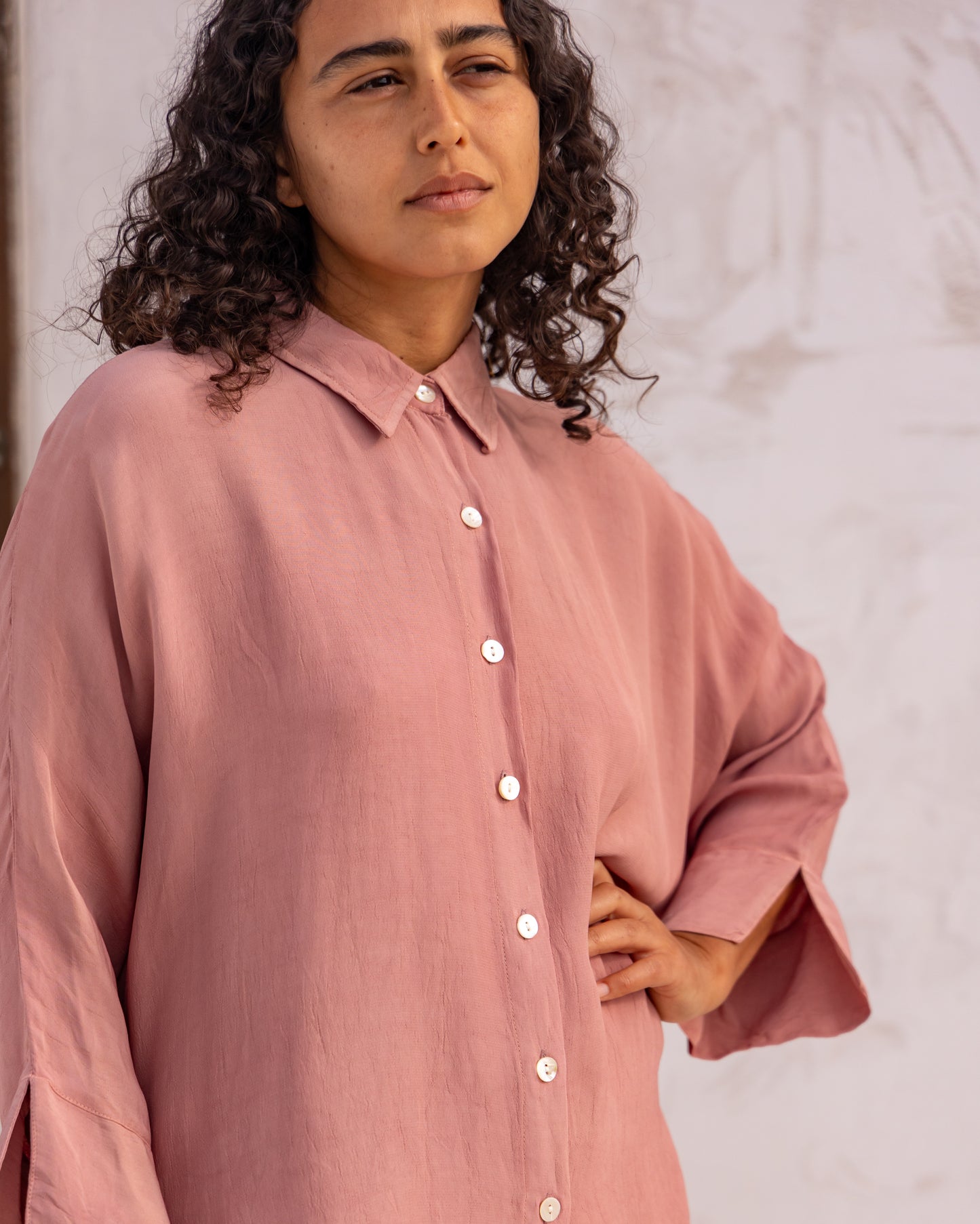 
                  
                    woman dressed in bouviet oversized shirt and miguel straight legged pants in orange peel fibre in dirty rose pink standing in front of a white wall
                  
                