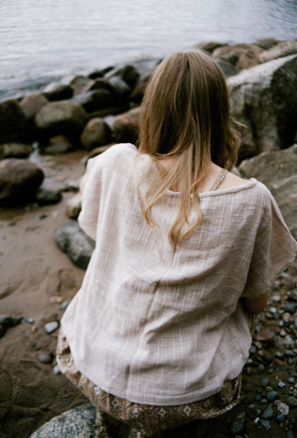 woman dressed in beige patterned mini dress with oversized net taupe top on rocky beach