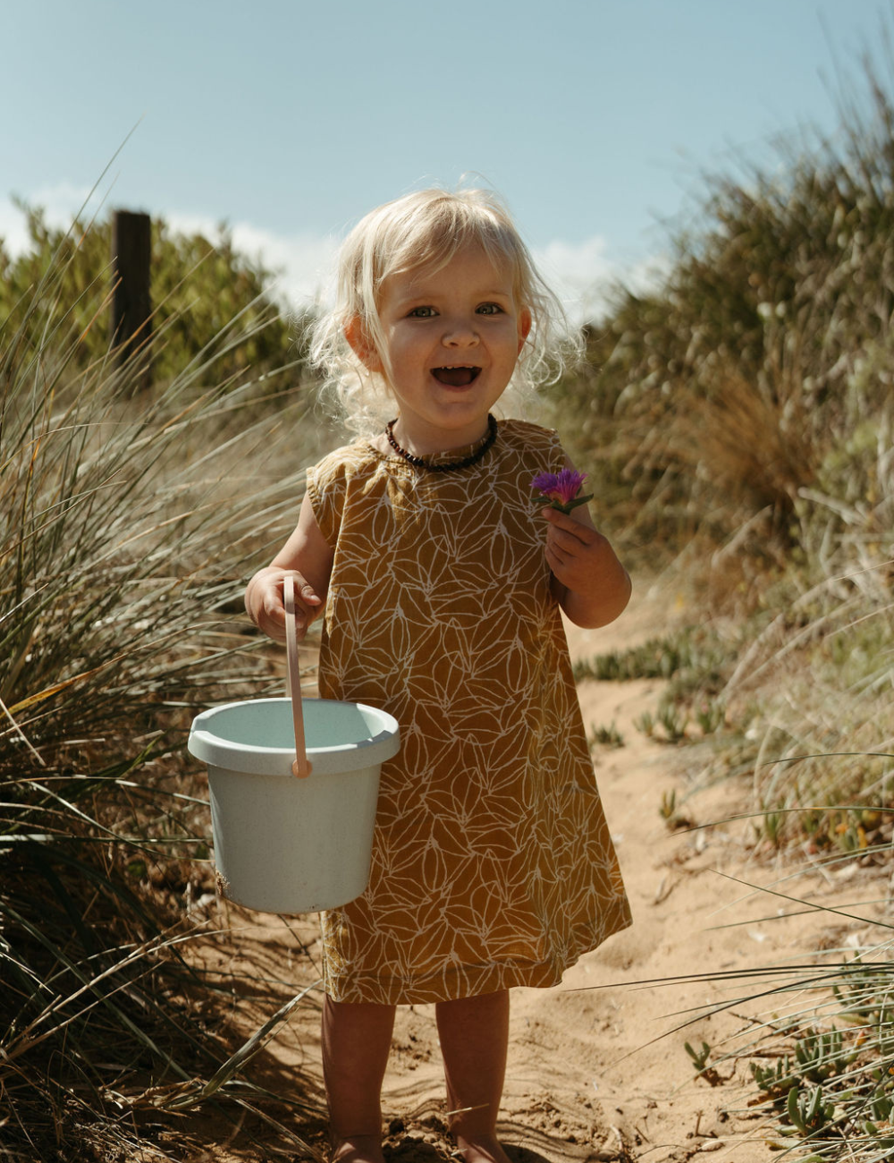 young girl with bucket and flower on beach in fogo straight dress in mustard seed print