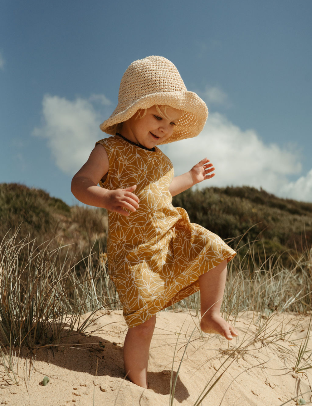 
                  
                     Young girl dancing on beach with a hat in fogo straight dress in mustard seed print
                  
                