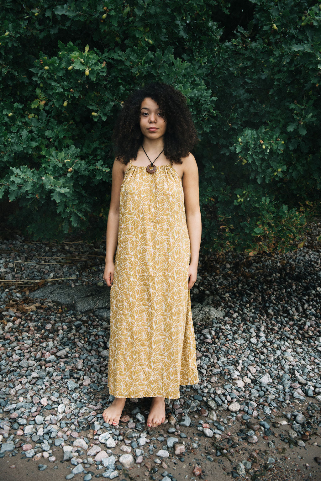 woman dressed in mustard and white patterned midi dress on rocky beach in front of trees