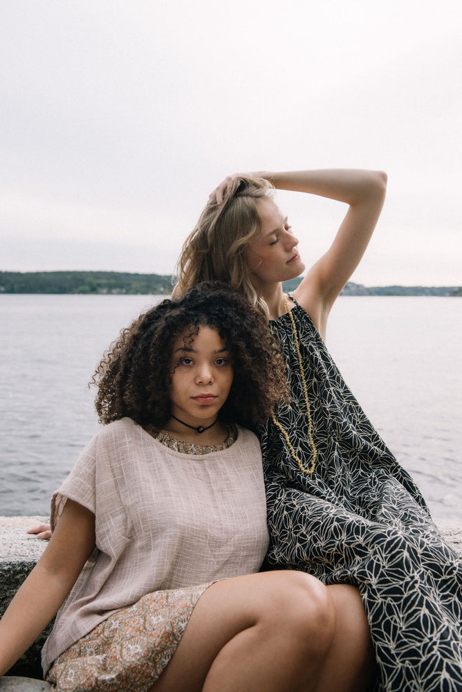 woman dressed in beige patterned mini dress with oversized net taupe top in front of ocean with another woman dressed in a dark patterned midi dress next to her
