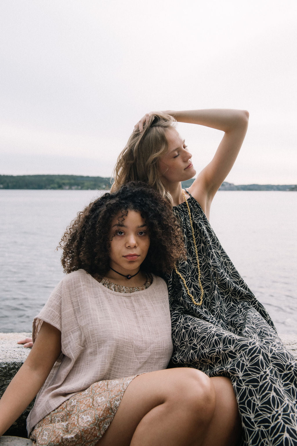 woman dressed in beige patterned mini dress with oversized net taupe top in front of ocean with another woman dressed in a dark patterned midi dress next to her