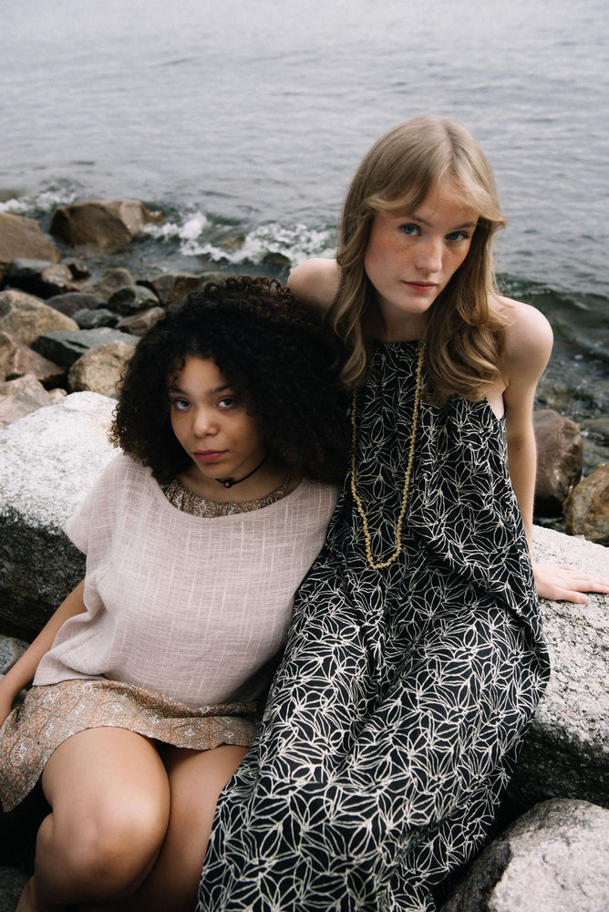 woman dressed in black and white patterned midi dress in front of rocks and ocean with another woman dressed in beige patterned mini dress and taupe coloured top sitting next to her