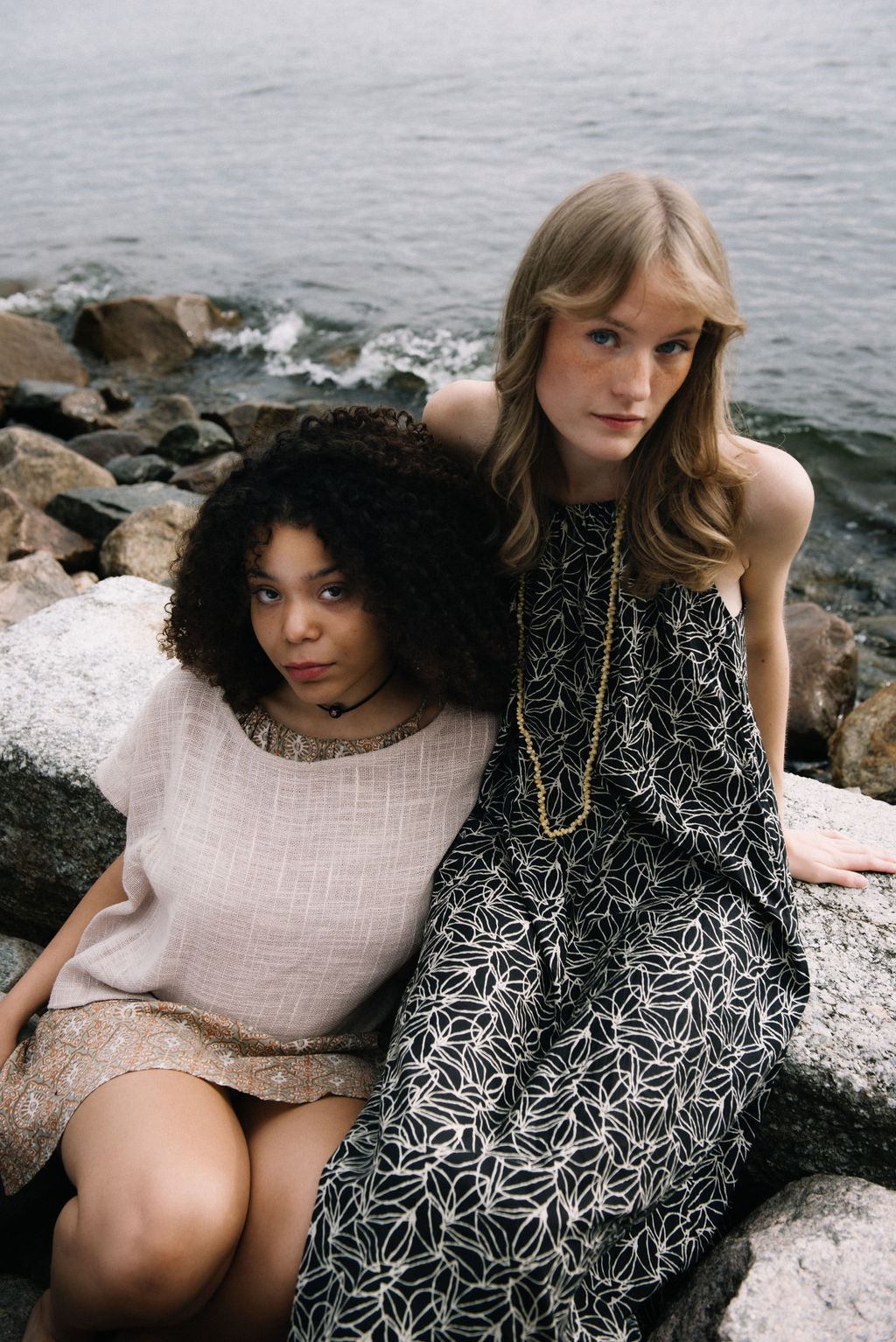 woman dressed in black and white patterned midi dress in front of rocks and ocean with another woman dressed in beige patterned mini dress and taupe coloured top sitting next to her