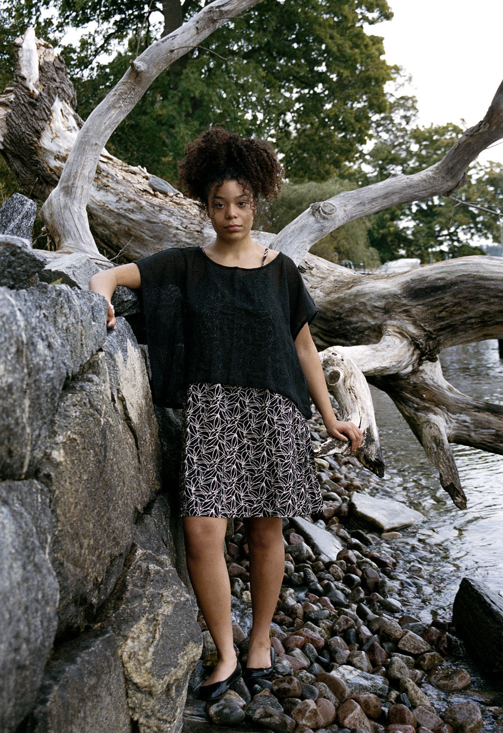 woman dressed in dark patterned mini dress with oversized net charcoal top in front of drift wood