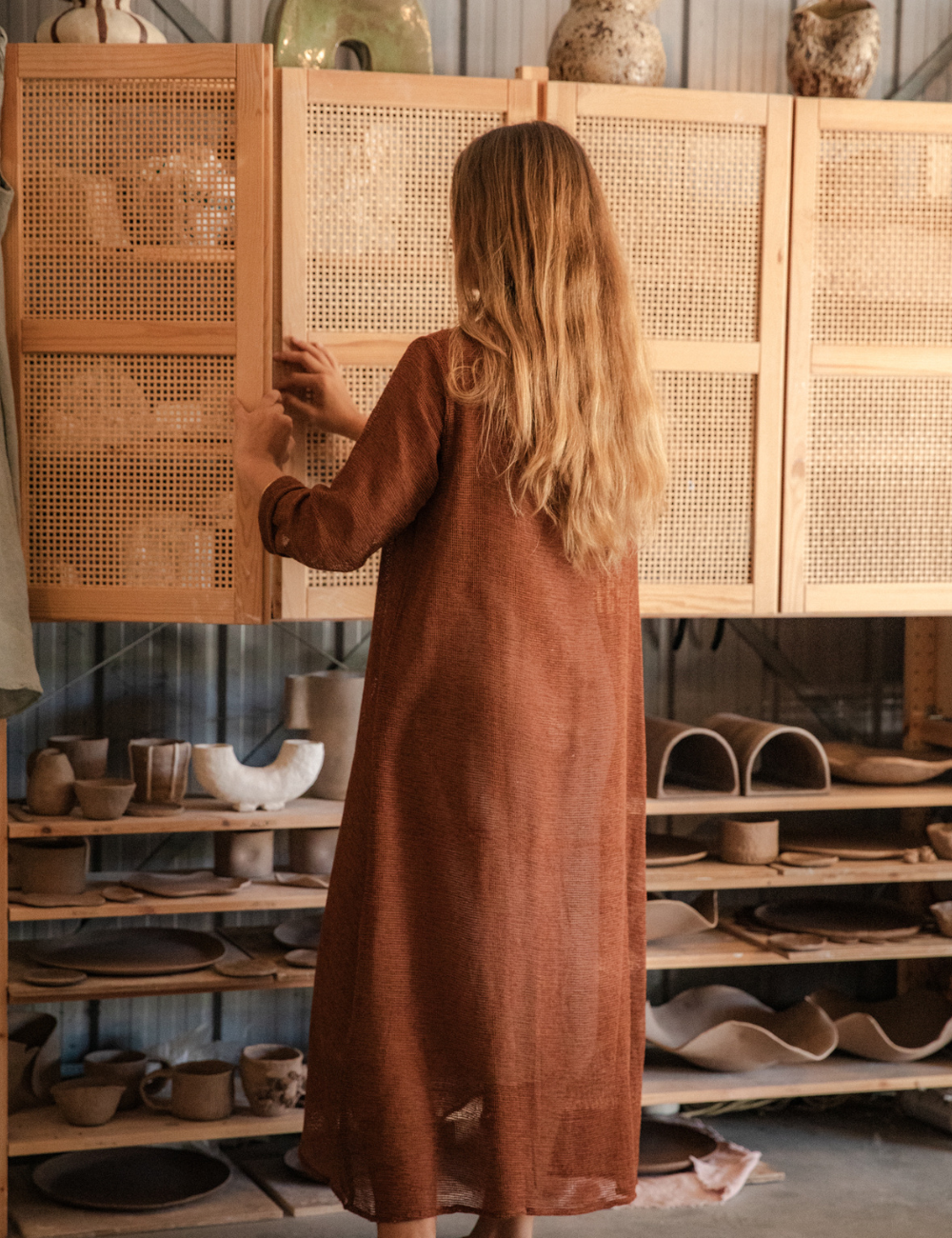 
                  
                    woman dressed in oversized maxi dress in net rust standing in pottery studio with ceramics in background
                  
                