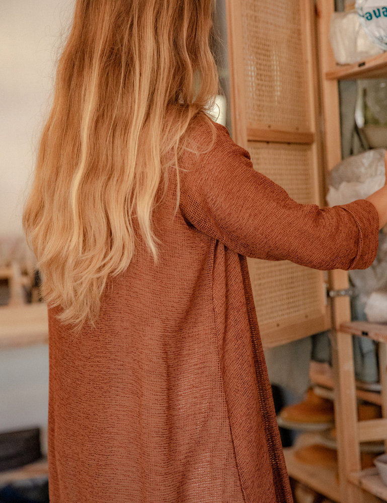 
                  
                    woman dressed in oversized maxi dress in net rust standing in pottery studio with ceramics in background
                  
                