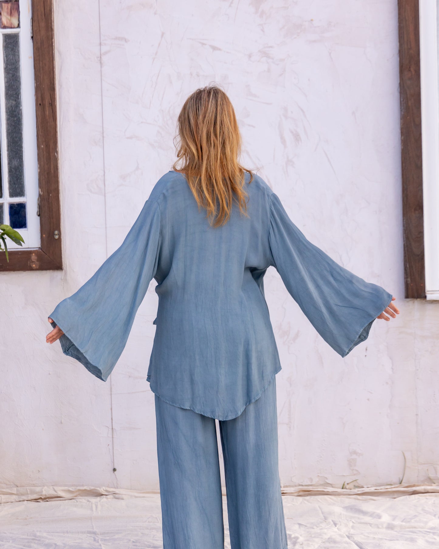 
                  
                    woman dressed in sao wrap top and miguel straight legged pants in orange peel fibre in blue cornflower standing in front of a white wall
                  
                
