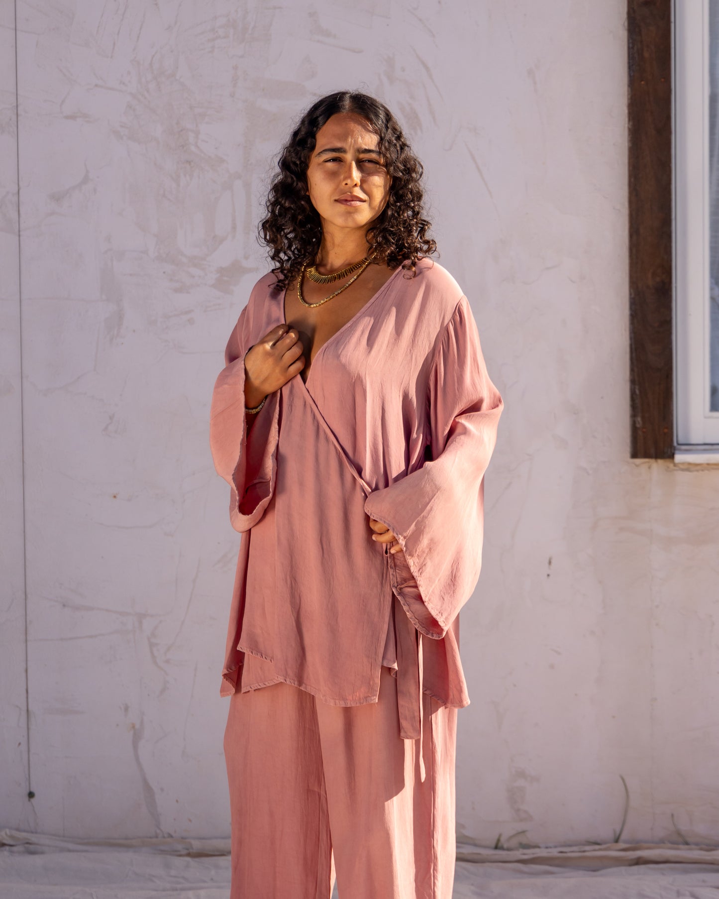
                  
                    woman dressed in sao wrap top and miguel straight legged pants in orange peel fibre in pink dirty rose standing in front of a white wall
                  
                