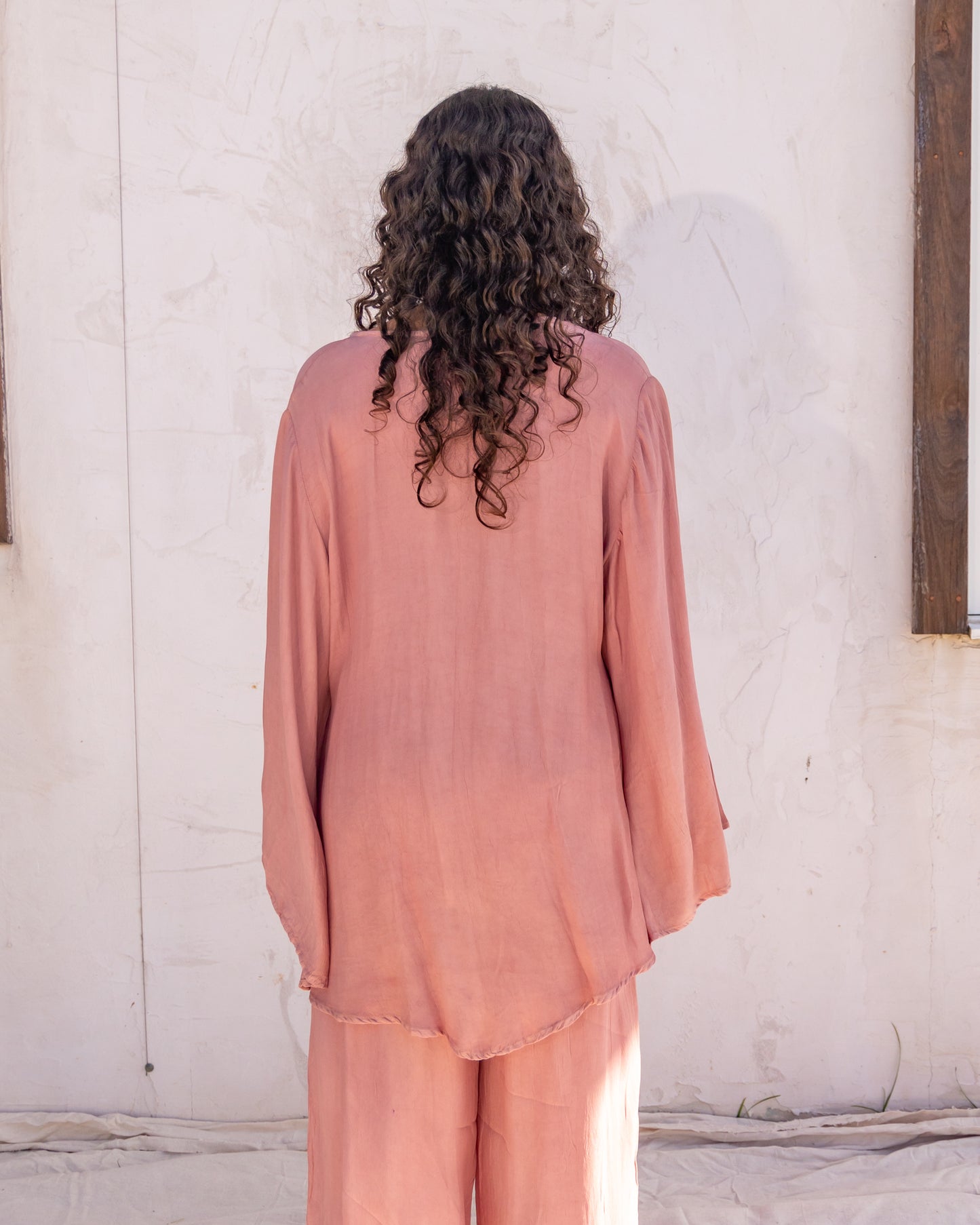 
                  
                    woman dressed in sao wrap top and miguel straight legged pants in orange peel fibre in pink dirty rose standing in front of a white wall
                  
                