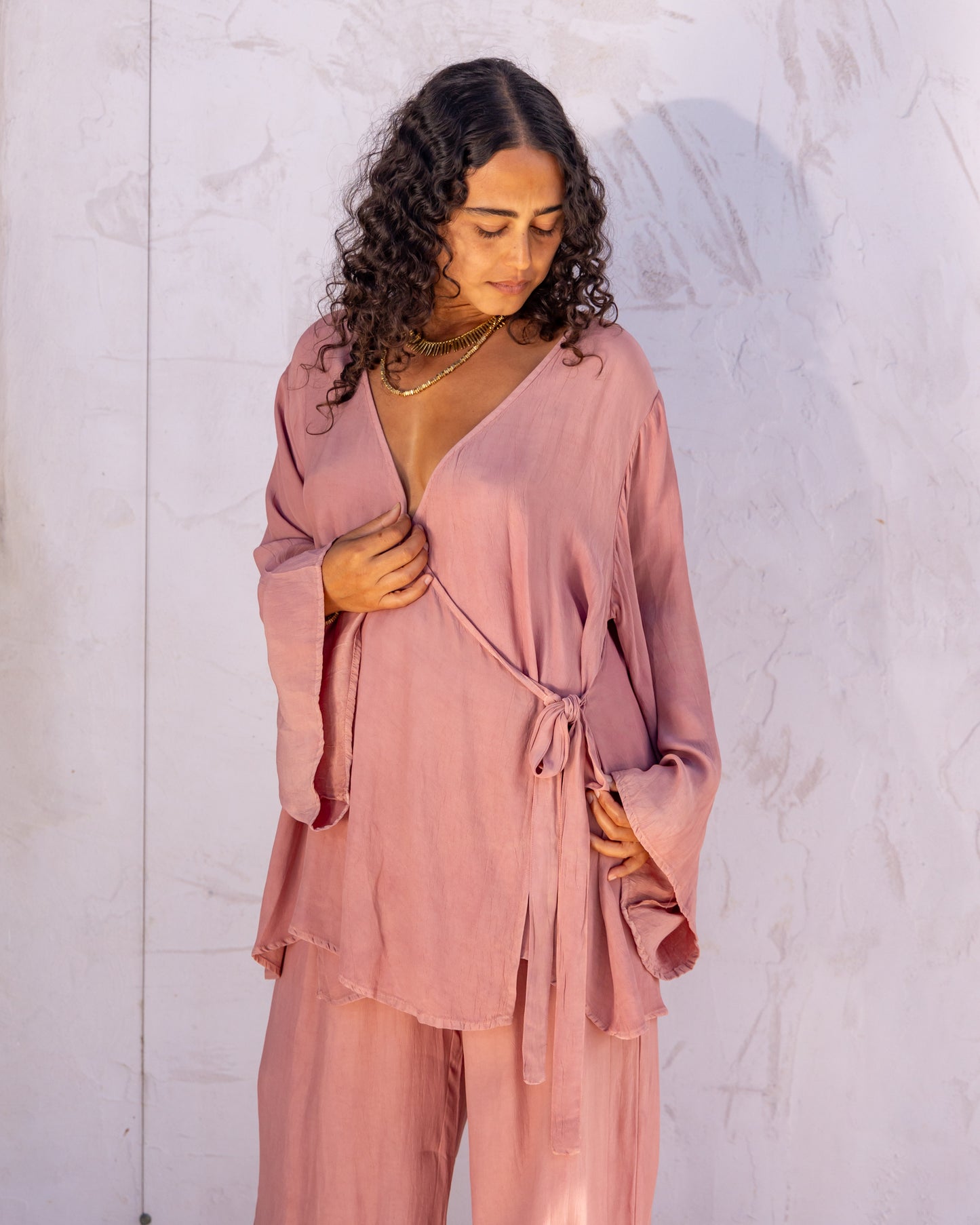 
                  
                    woman dressed in sao wrap top and miguel straight legged pants in orange peel fibre in pink dirty rose standing in front of a white wall
                  
                