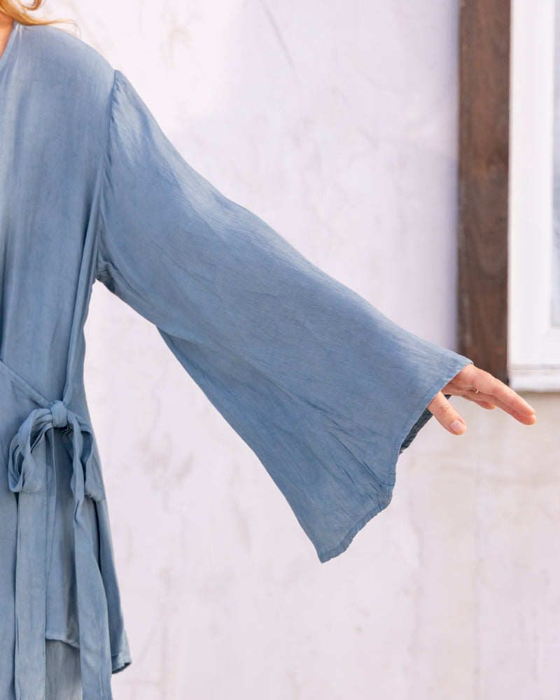 
                  
                    woman dressed in sao wrap top and miguel straight legged pants in orange peel fibre in blue cornflower standing in front of a white wall
                  
                