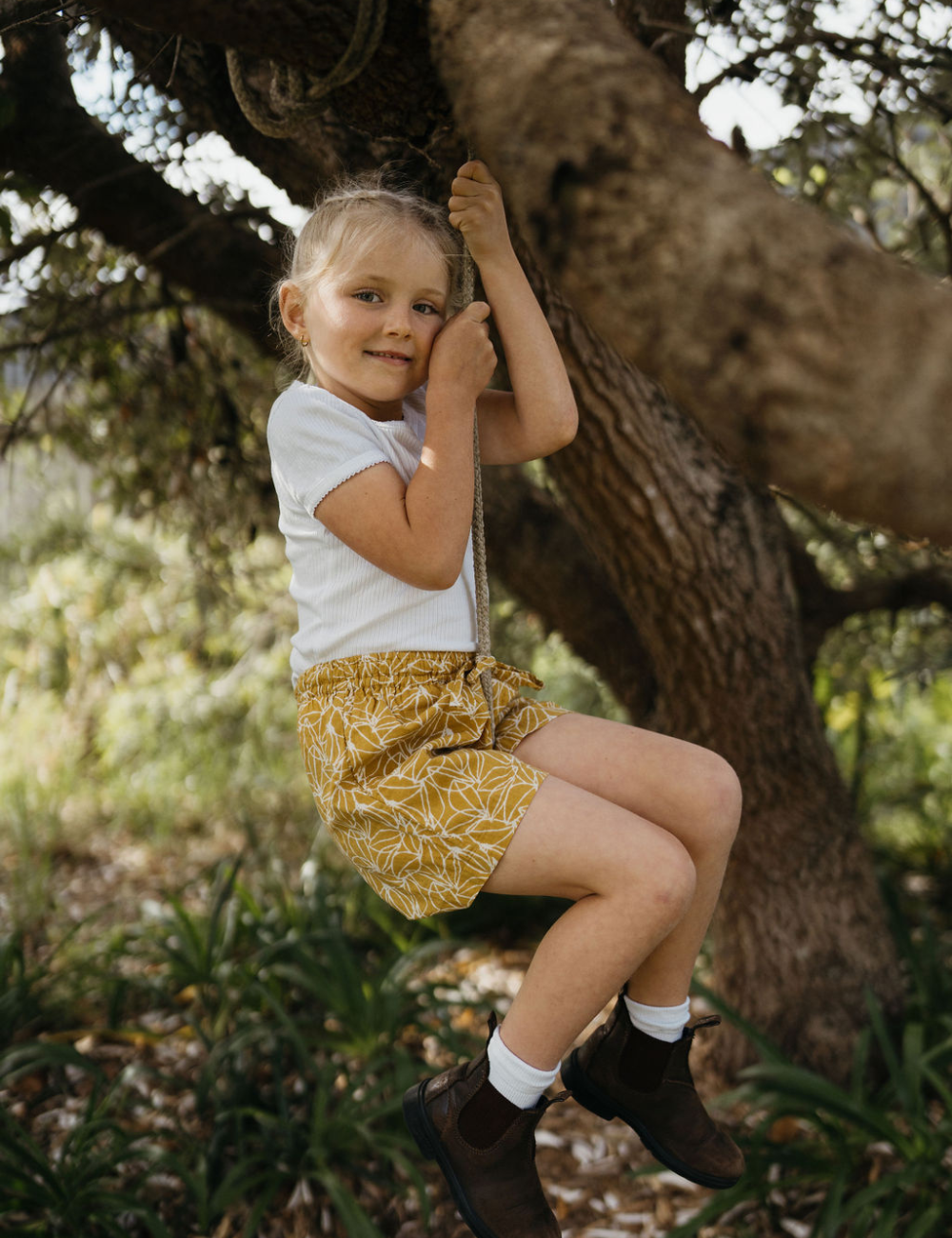 
                  
                    young girl swinging from a tree dressed in white singlet and retro block printed shorts in mustard seed print
                  
                