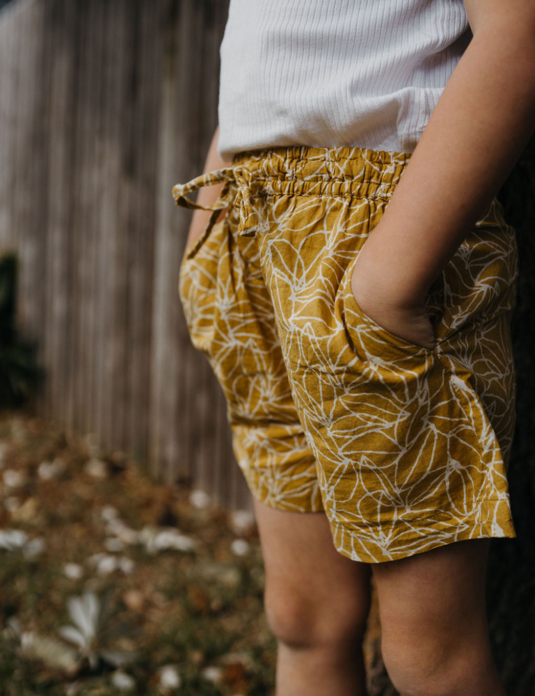 
                  
                    young girl in front of fence dressed in white singlet and retro block printed shorts in mustard seed print
                  
                