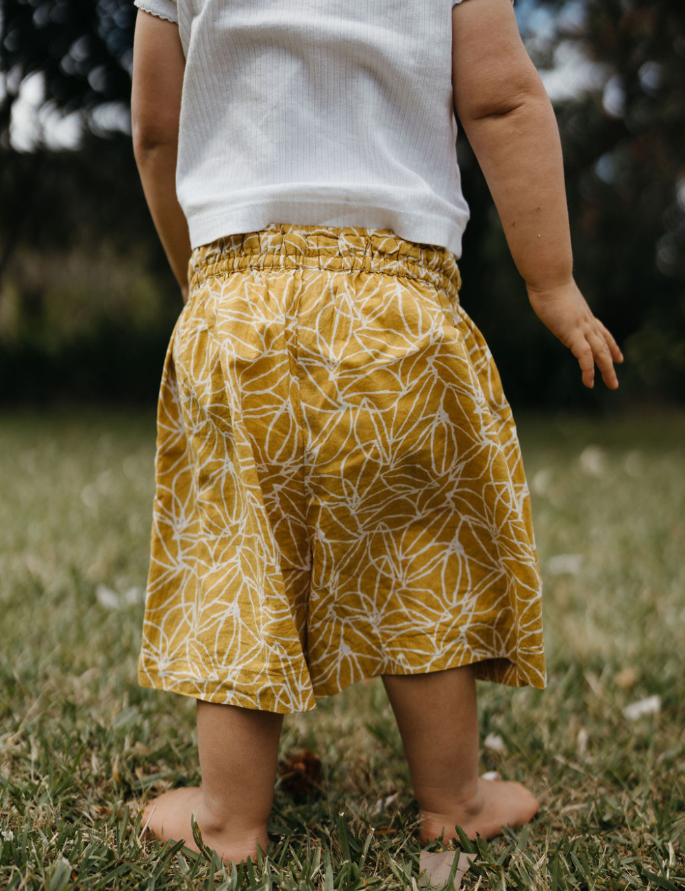 young child from behind on lawn dressed in white singlet and retro block printed shorts in mustard seed print