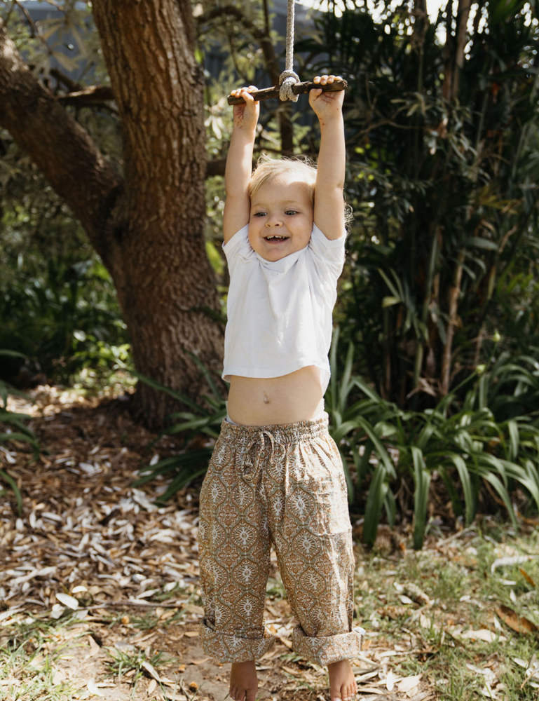 
                  
                    young child dressed in white singlet and Indian retro block printed pants in retro funk print hanging on a swing in forest
                  
                